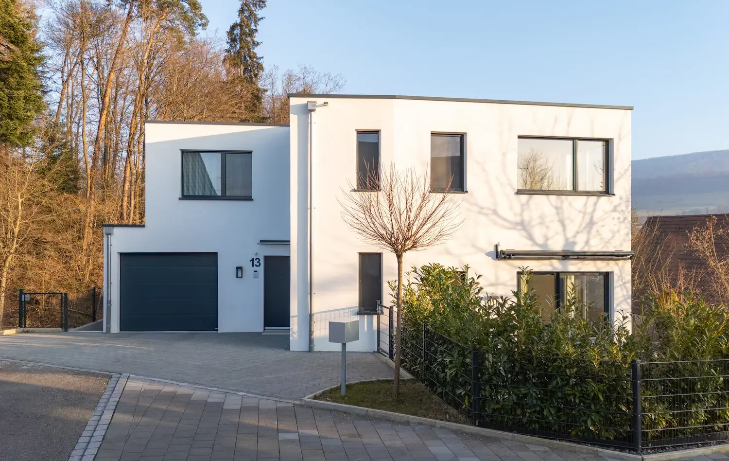Neubau eines modernen Einfamilienhauses mit weisser Fassade, Flachdach und kubischer Architektur. Zweigeschossiges Wohnhaus mit Untergeschoss, integrierter Garage, grossen Fensterflächen und gepflegtem Aussenbereich in ruhiger Wohnlage mit Bäumen im Hintergrund.