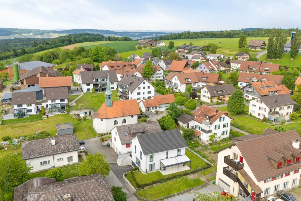 Luftaufnahme eines ländlichen Wohngebiets mit Einfamilienhäusern und Kirche, eingebettet in grüne Wiesen und Hügel. Ruhige Wohnlage in naturnaher Umgebung.