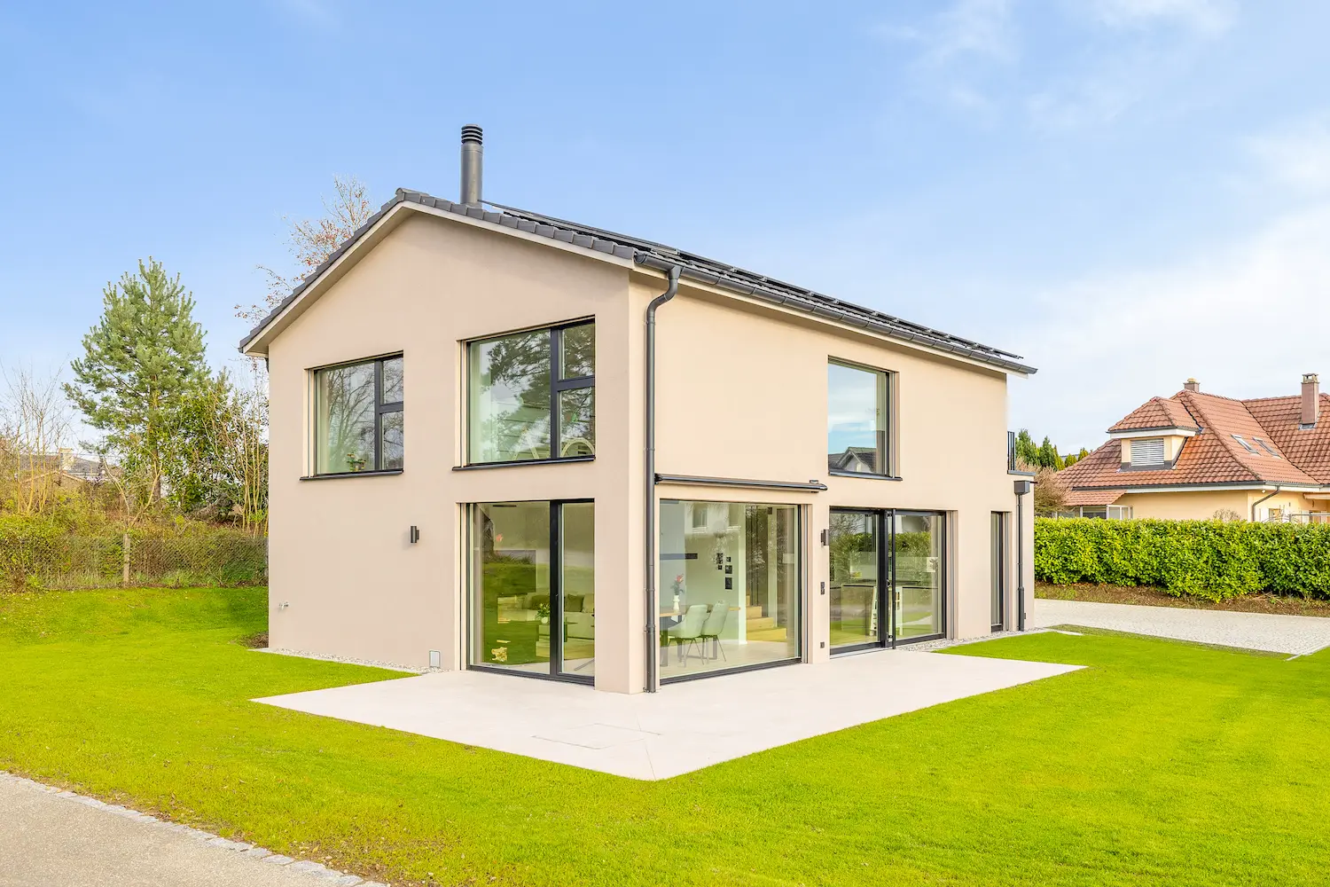 Modernes Einfamilienhaus mit warmer, beige verputzter Fassade, klarer Architektur, grossen Fensterflaechen, flachem Satteldach und gepflegtem Garten in ruhiger Wohnlage.