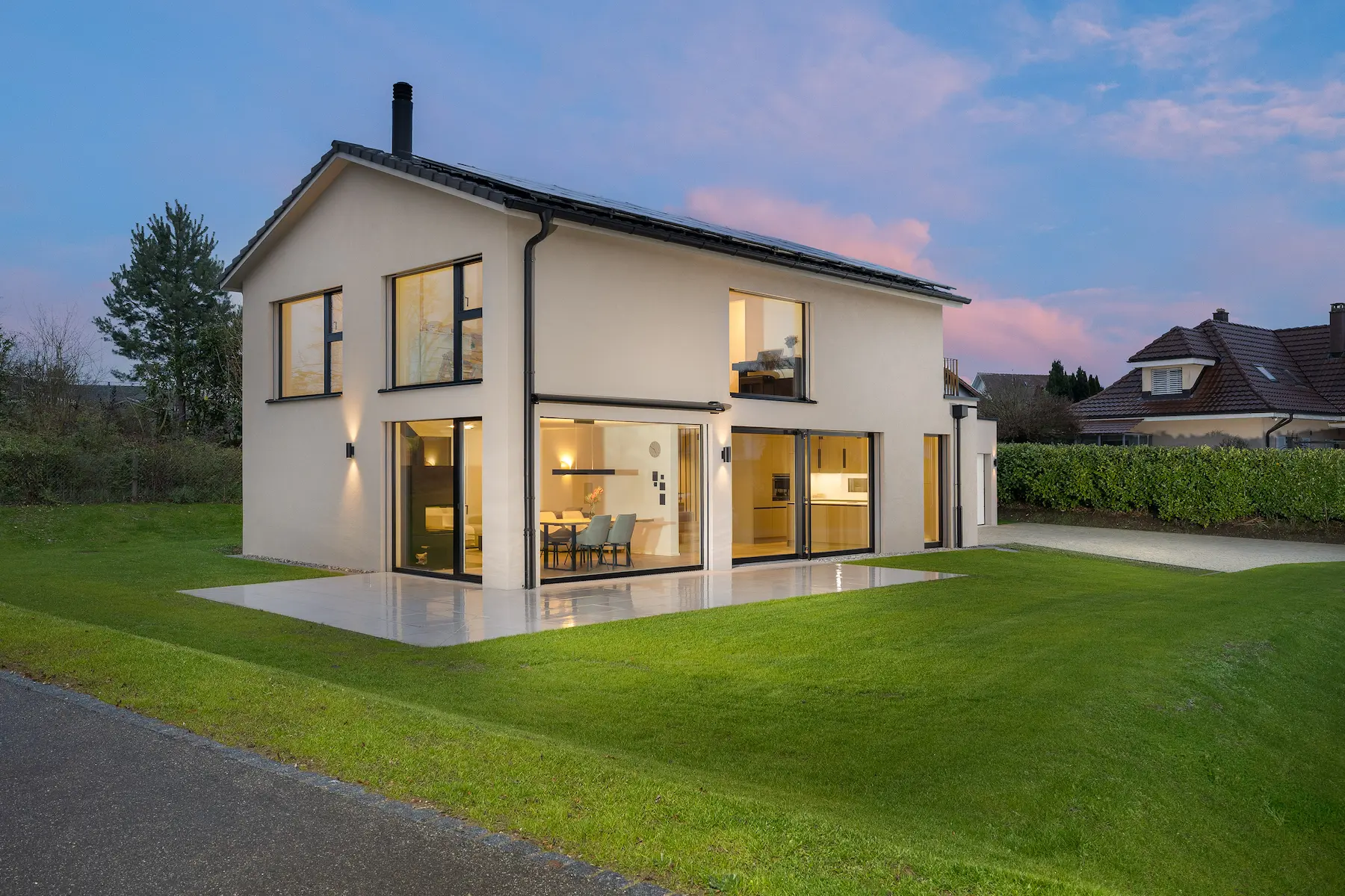Modernes Einfamilienhaus mit heller Fassade, grosser Glasfront, Terrasse und gepflegtem Garten bei Abenddämmerung.