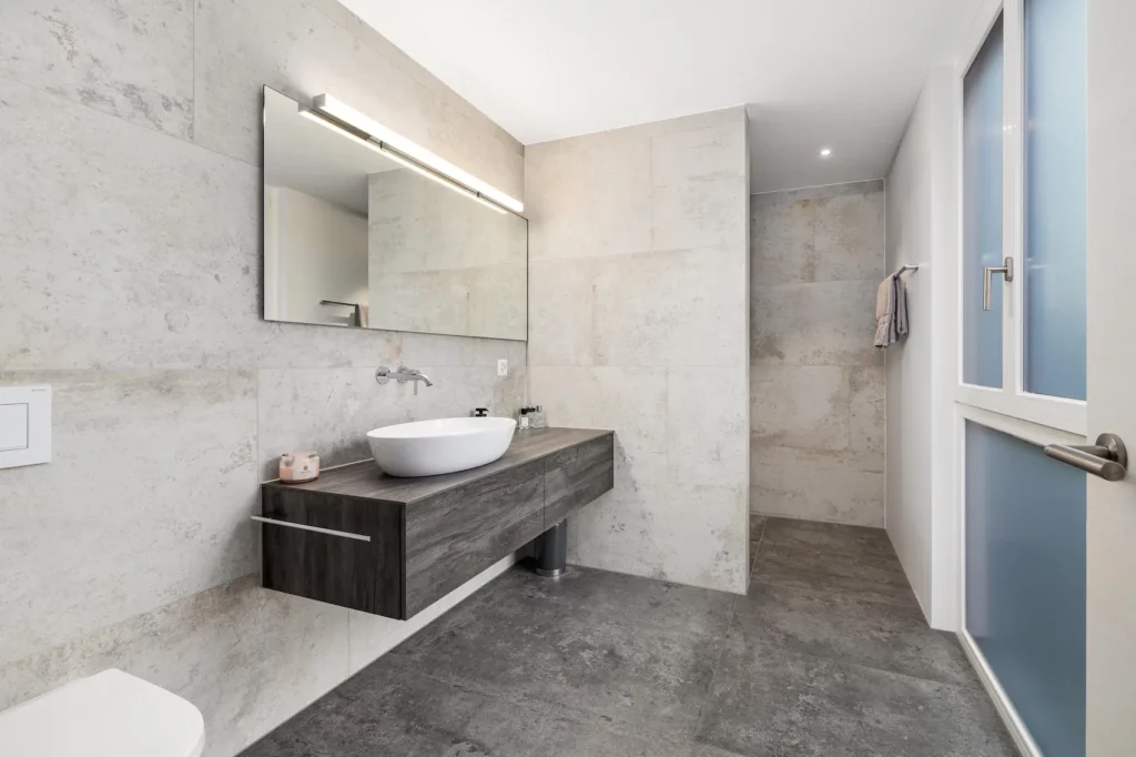 Modernes, helles Badezimmer mit grossformatigen Steinfliesen, bodengleicher Dusche und klaren Linien für eine ruhige, zeitlose Raumwirkung.