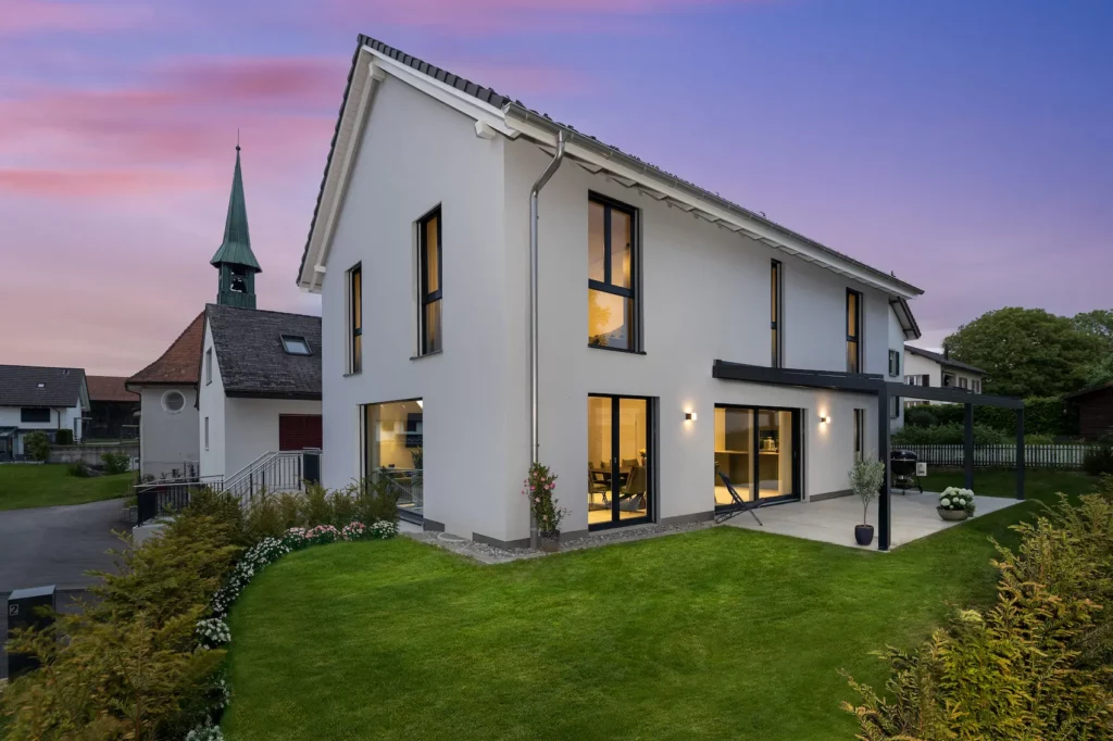 Modernes Einfamilienhaus in Massivbauweise bei Dämmerung mit grossen Fensterflächen, Satteldach, gepflegtem Garten und zeitgemässer Architektur.