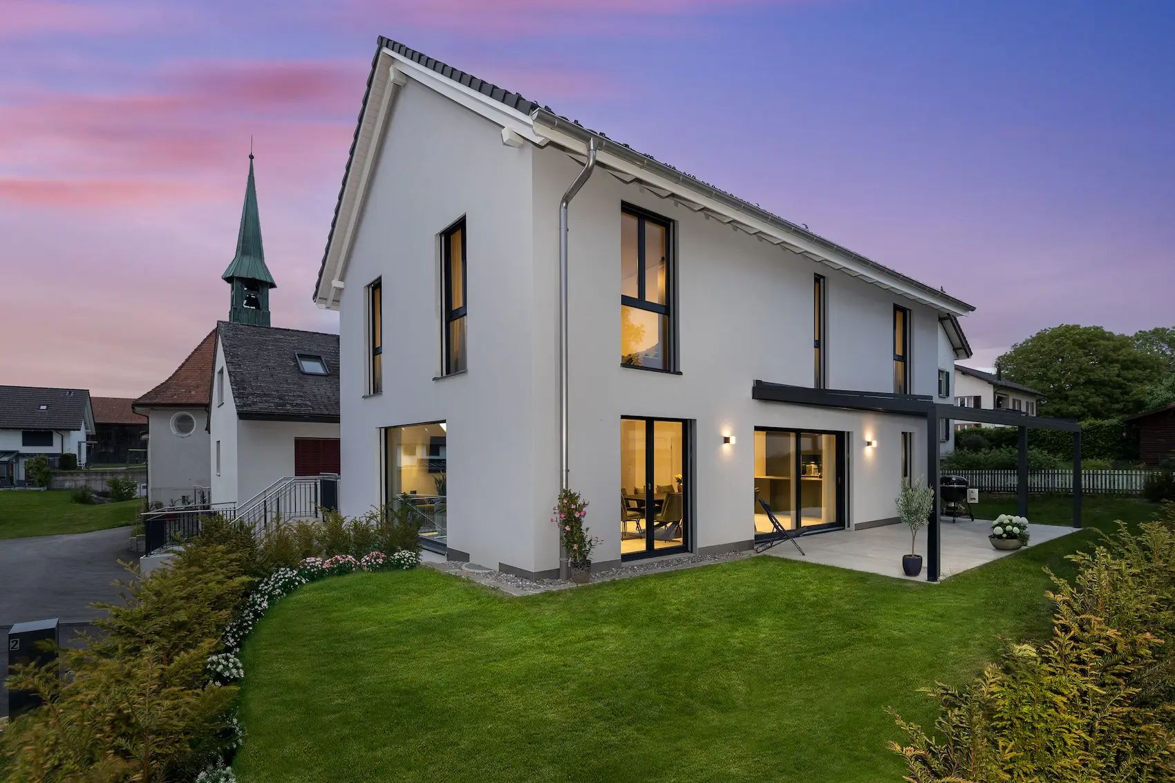 Modernes Einfamilienhaus in Massivbauweise bei Dämmerung mit grossen Fensterflächen, Satteldach, gepflegtem Garten und zeitgemässer Architektur.