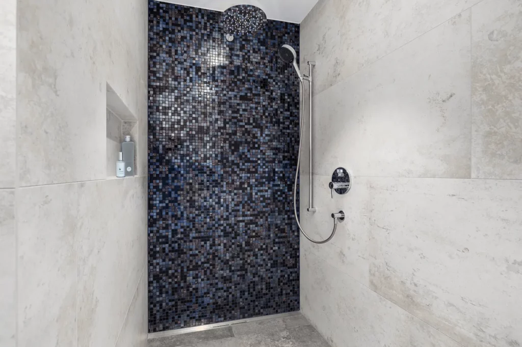 Bodengleiche Dusche mit hellen Natursteinfliesen, dunkler Mosaik-Akzentwand, Regenduschkopf und integrierter Wandnische in modernem Badezimmer.