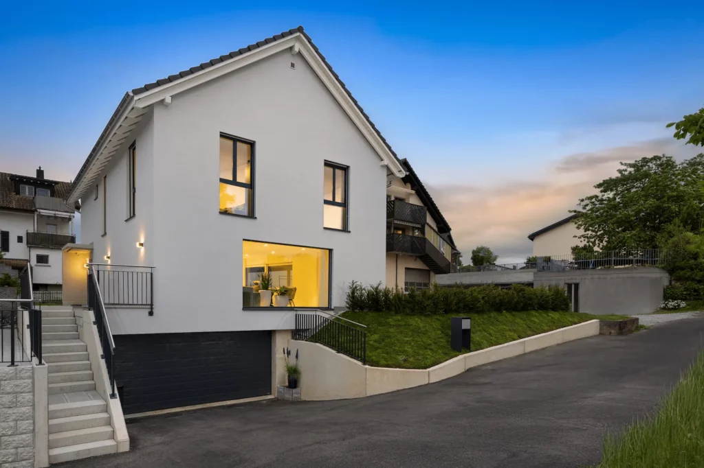 Modernes Einfamilienhaus mit weißer Fassade und Satteldach, Abendstimmung mit beleuchteten Wohnräumen und Hanglage.