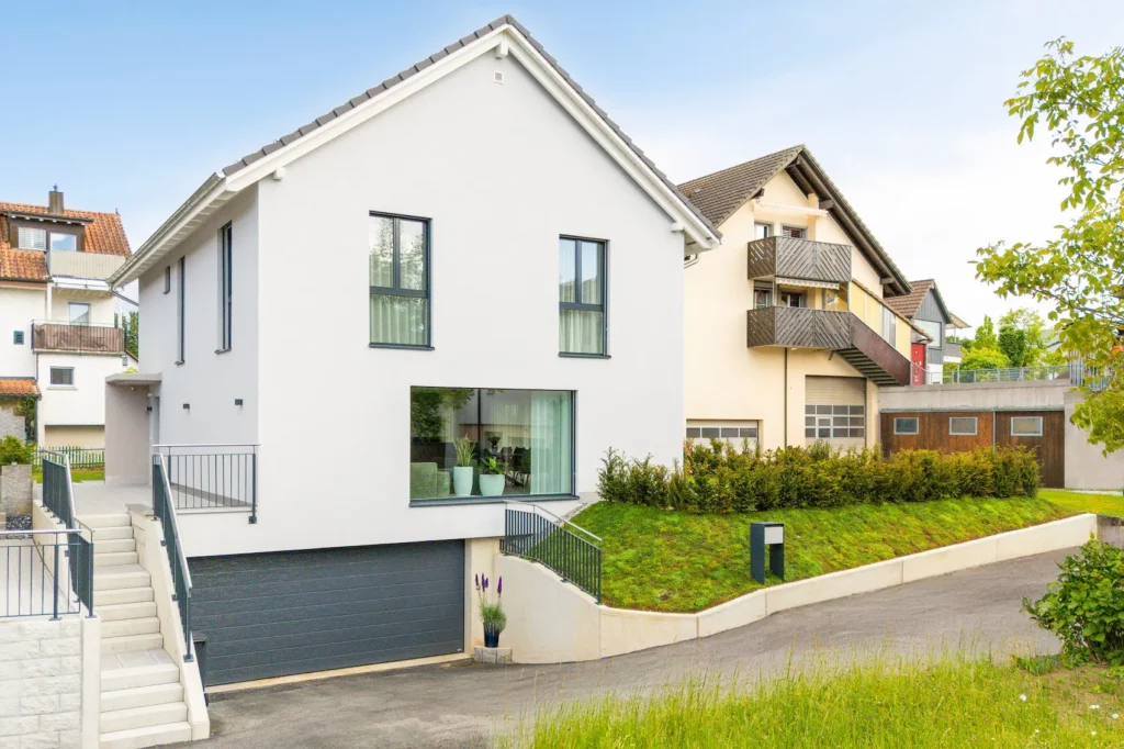 Modernes Einfamilienhaus mit heller Fassade, klarer Architektur, integrierter Garage und leichter Hanglage in ruhigem Wohnquartier.