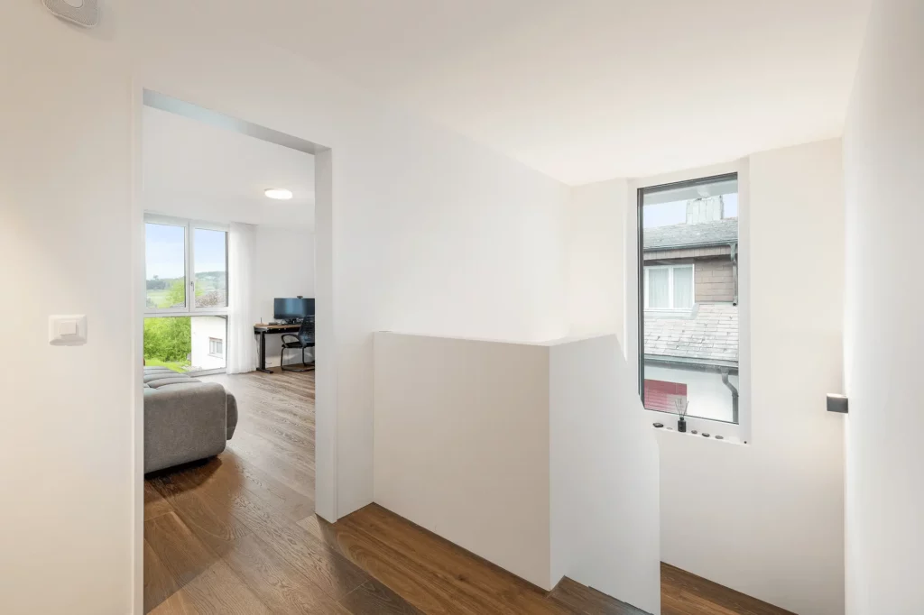 Heller, minimalistischer Flur im Obergeschoss mit weiß gestrichenen Wänden, Holzparkettboden und Fenster; Blick durch eine offene Tür in ein Arbeitszimmer mit Schreibtisch und Monitor.