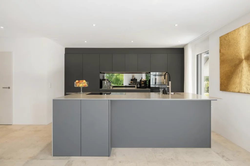 Offene Designküche mit großer grauer Kochinsel, matten Fronten, schwarzer Armatur und integrierten Geräten, minimalistisch gestaltet und durch Tageslicht hell ausgeleuchtet.