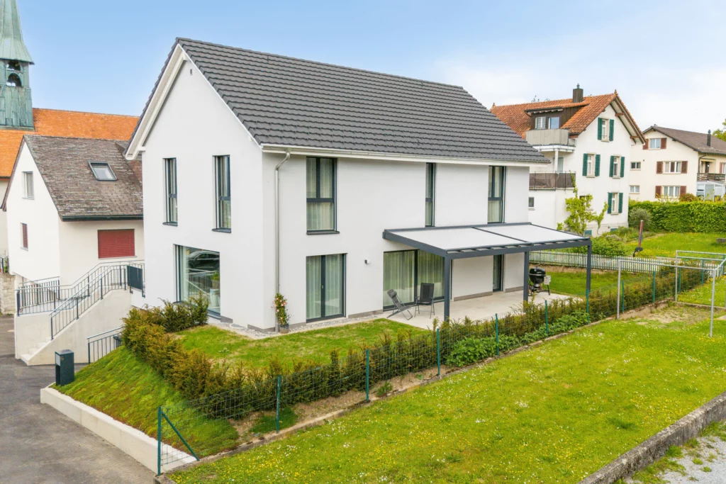 Modernes Einfamilienhaus mit weißer Fassade und Satteldach, großzügiger überdachter Terrasse und gepflegtem Garten in ruhigem Wohnquartier.