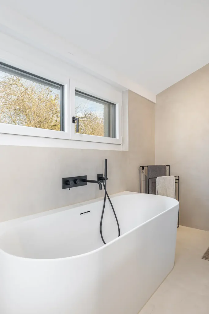 Freistehende Badewanne mit schwarzer Armatur vor Fenster, in hell gestaltetem Badezimmer.