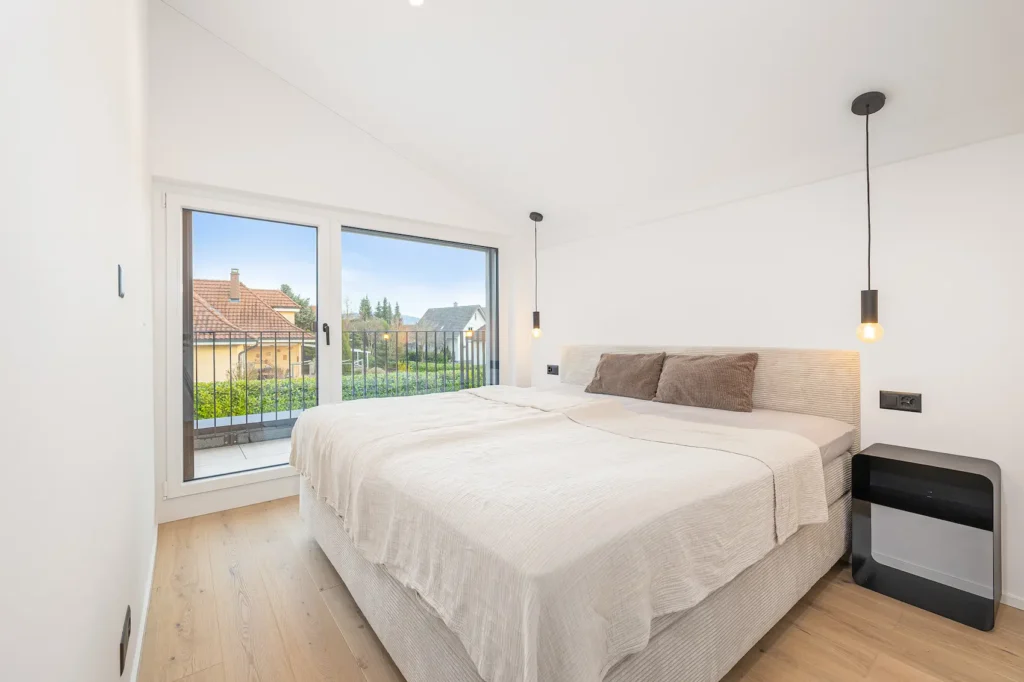 Helles Schlafzimmer mit Doppelbett, bodentiefem Fenster, Zugang zum Balkon und Pendelleuchten neben dem Bett.