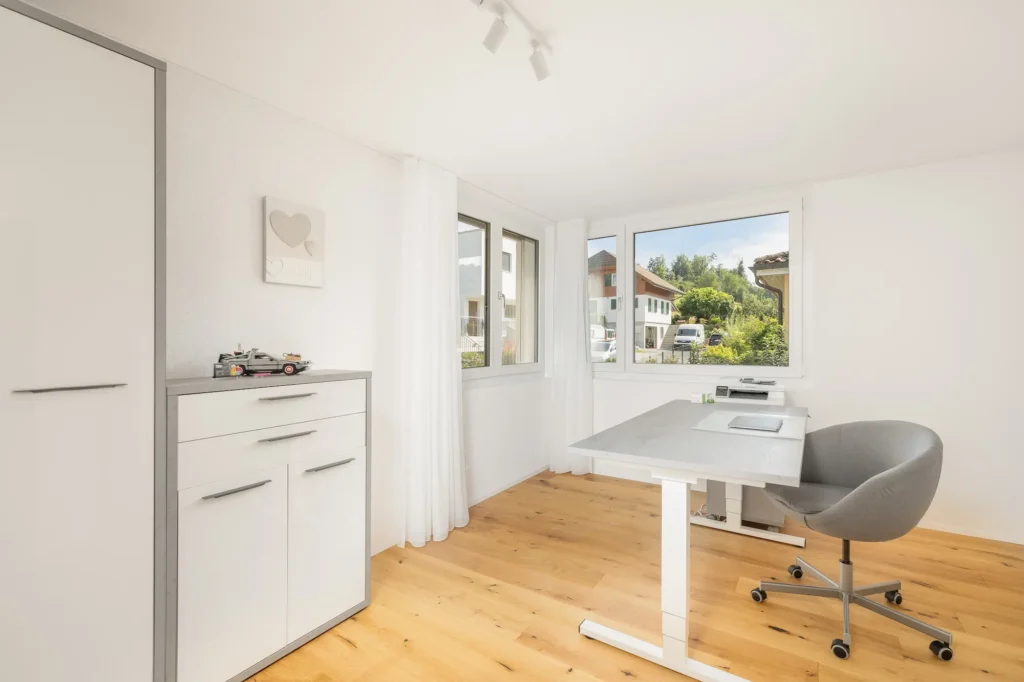 Helles Arbeitszimmer mit Schreibtisch, Schrankelementen, Vorhängen und grosszügiger Fensterfläche.