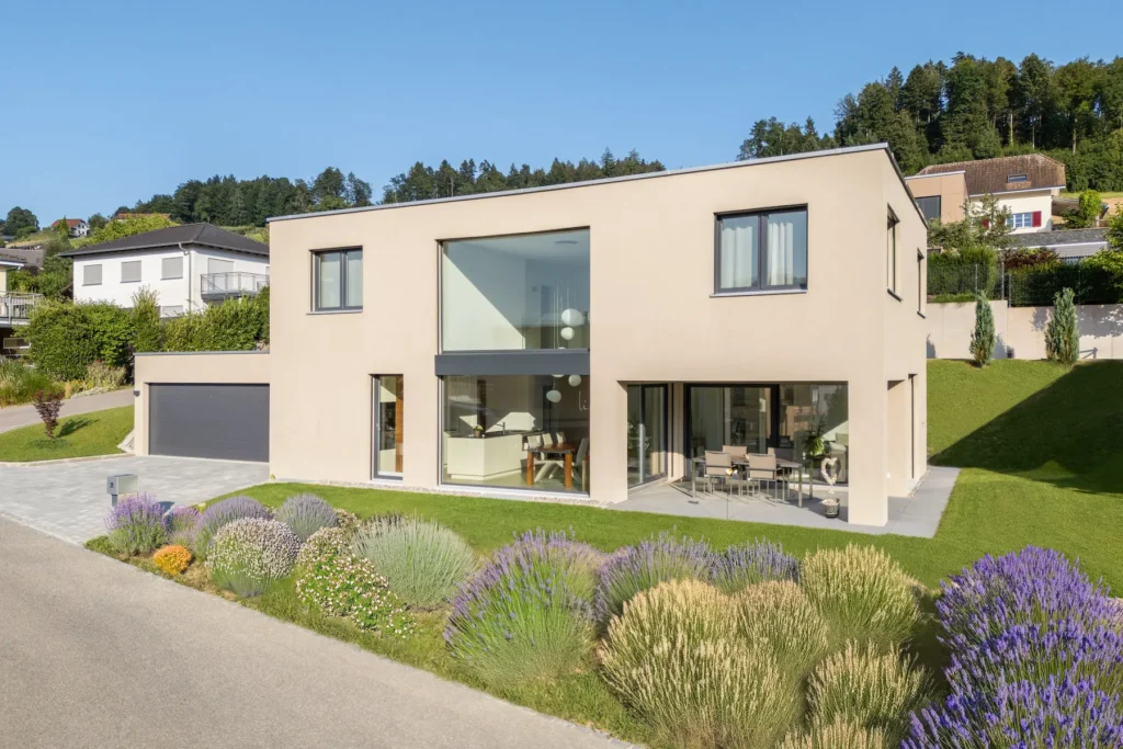 Aussenansicht eines modernen Einfamilienhauses mit Flachdach, grossen Fensterflächen, Terrasse und gestalteter Gartenanlage.