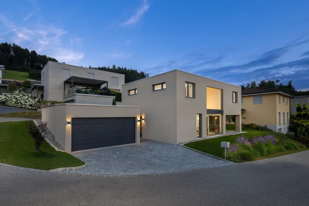 Aussenansicht eines modernen Einfamilienhauses mit Flachdach in der Dämmerung mit beleuchteter Garage, Vorplatz und Fensterflächen.