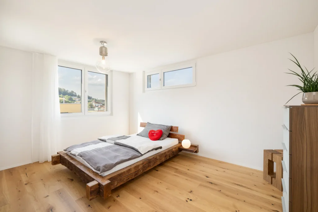 Helles Schlafzimmer mit massivem Holzbett, Holzparkett, grossen Fenstern und minimaler Einrichtung.