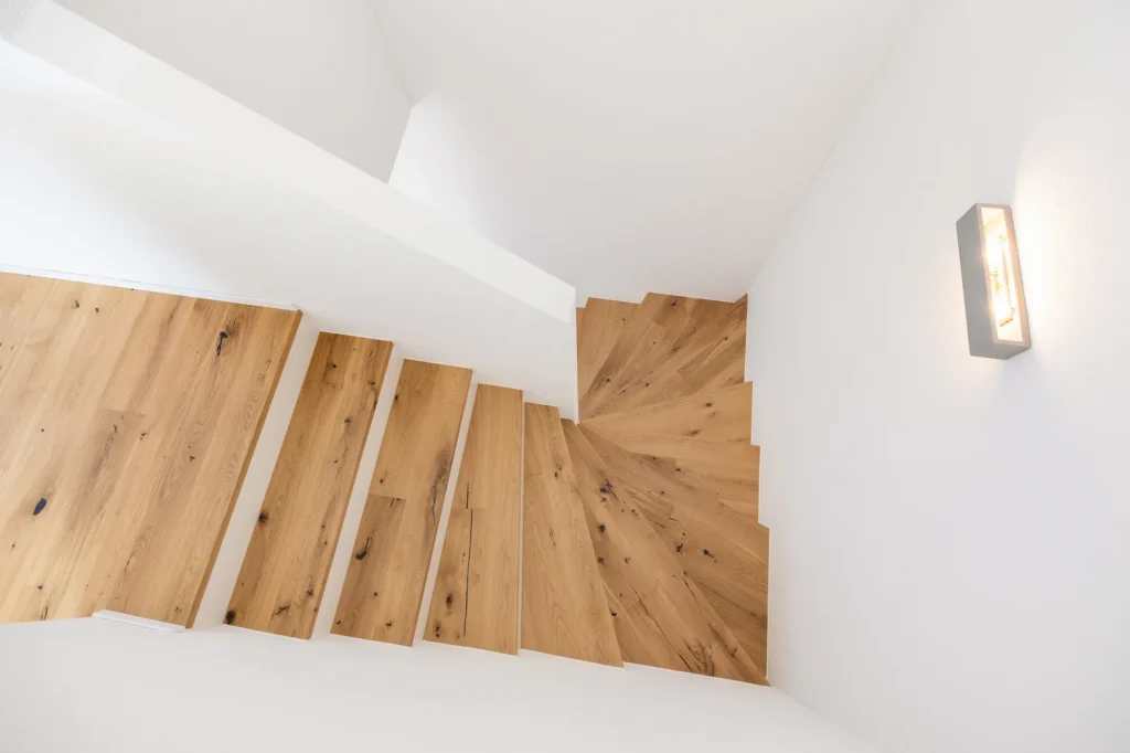Treppenaufgang mit massiven Holzstufen, gewendeter Geometrie, weissen Wänden und Wandleuchte.