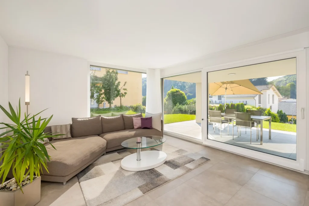Wohnzimmer mit Ecksofa, bodentiefen Fenstern und Schiebetür zur überdachten Terrasse und zum Garten.
