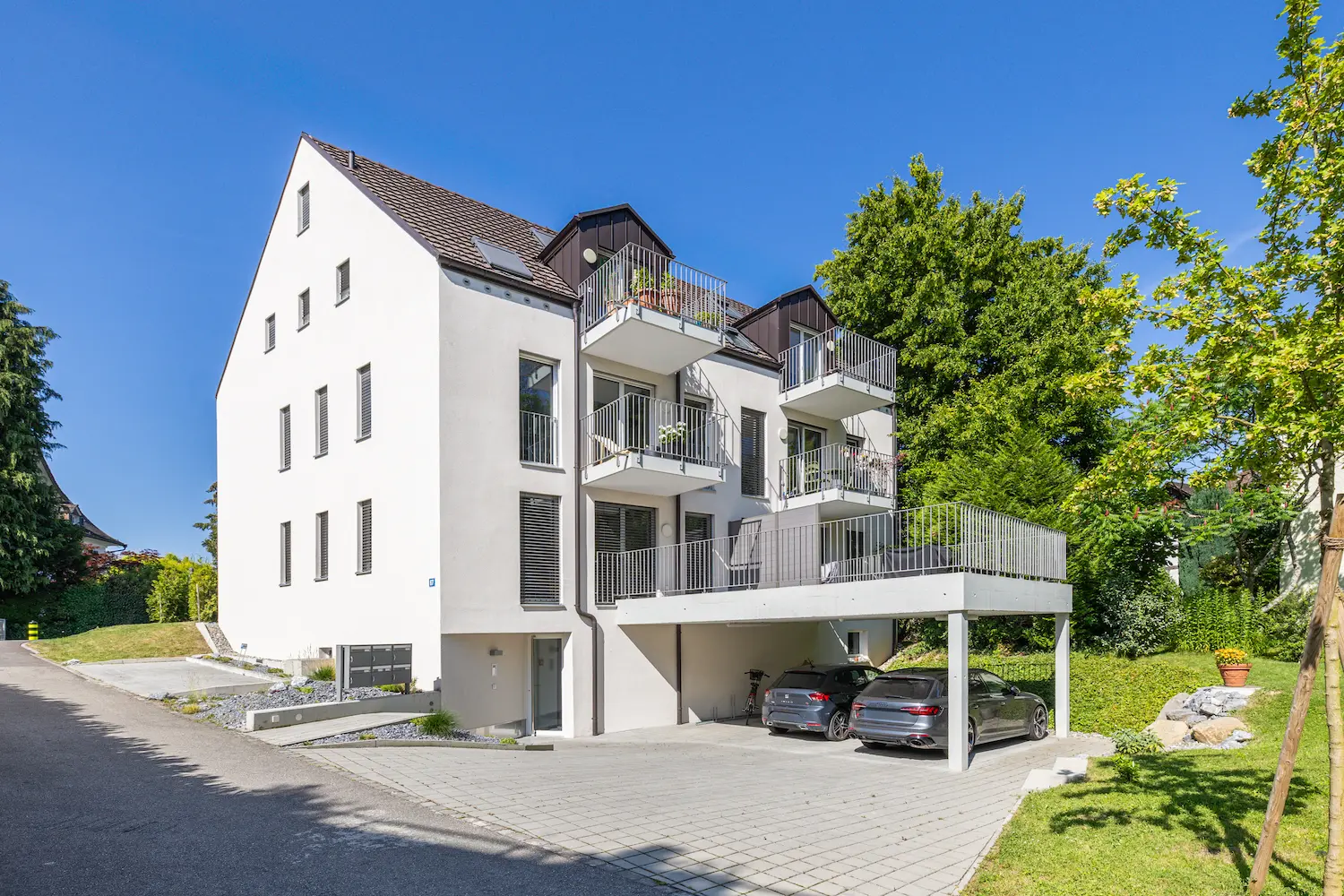 Modernes Mehrfamilienhaus mit heller, weißer Fassade, Balkonen und überdachtem Parkplatz umgeben von Grün in städtischer Lage.