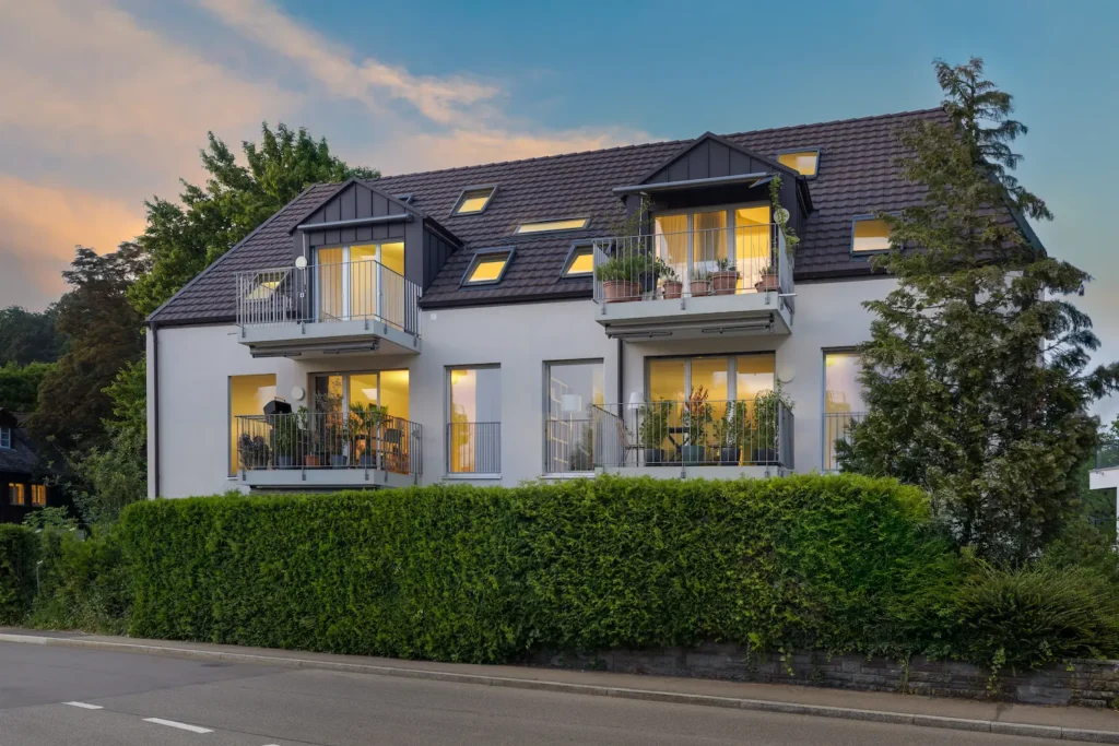 Modernes Mehrfamilienhaus mit mehreren Balkonen und beleuchteten Wohnungen in der Abenddämmerung in Zürich.