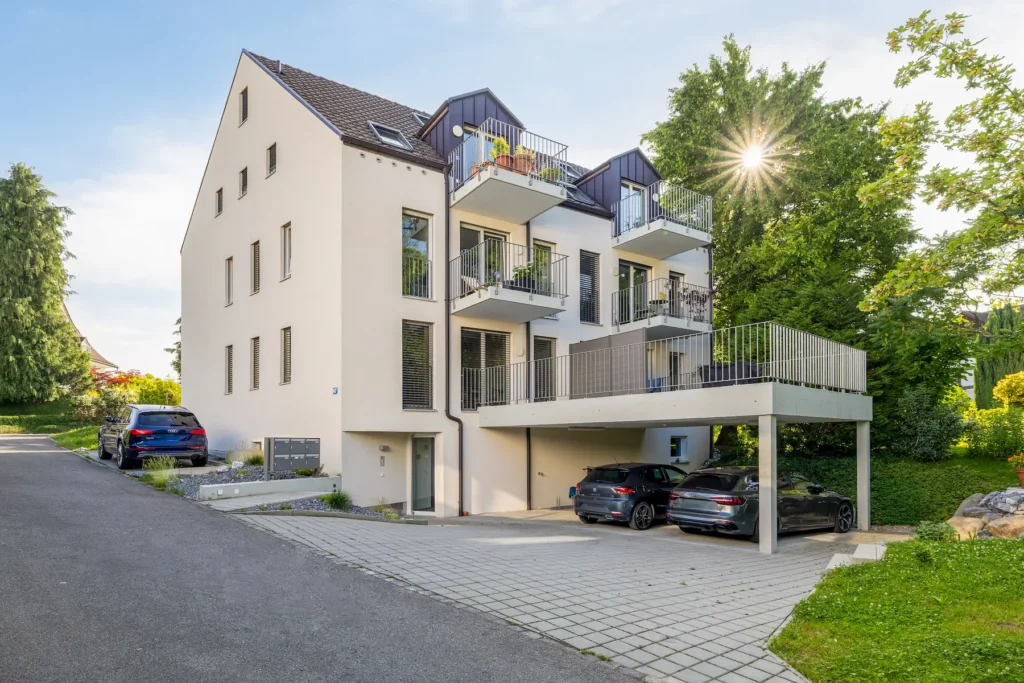 Mehrfamilienhaus in Zürich mit seitlichem Haupteingang, Balkonen und überdachten Parkplätzen im Grünen.
