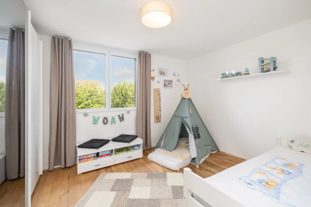 Heller Raum mit Fenster, Holzboden, Bett und Spielbereich.
