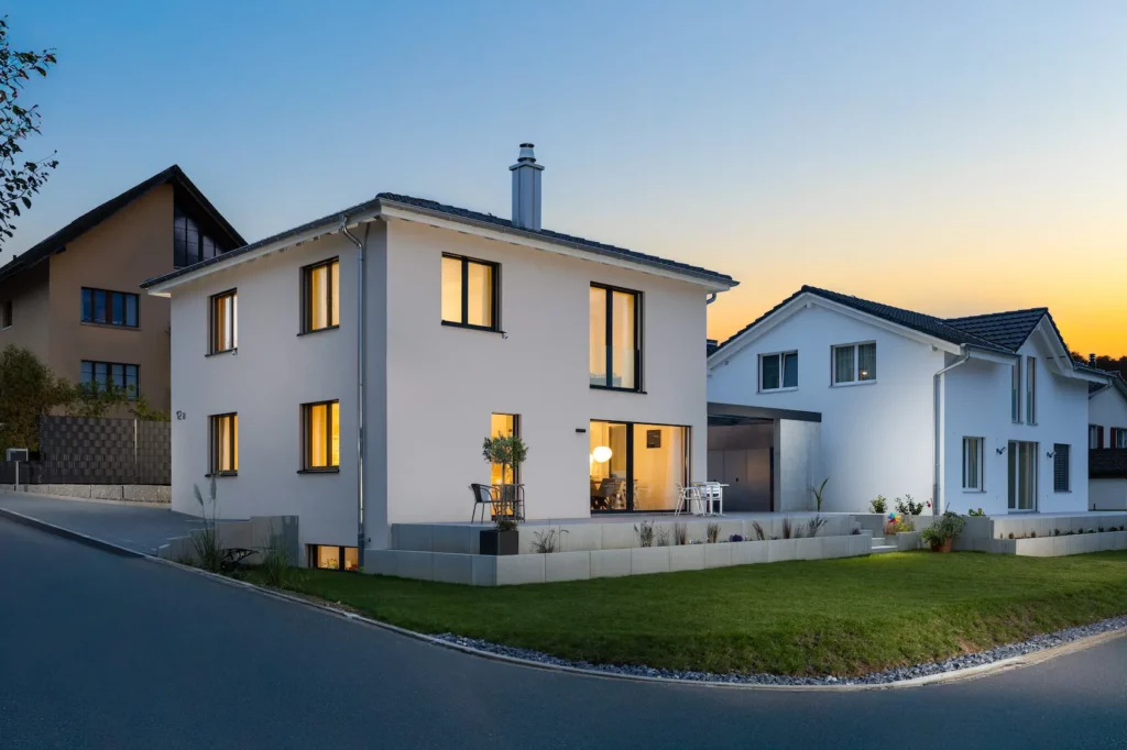 Modernes Einfamilienhaus mit heller, weiss verputzter Fassade, klarer Architektur, grossen Fenstern und Garten in ruhiger Wohnlage bei Abendstimmung.