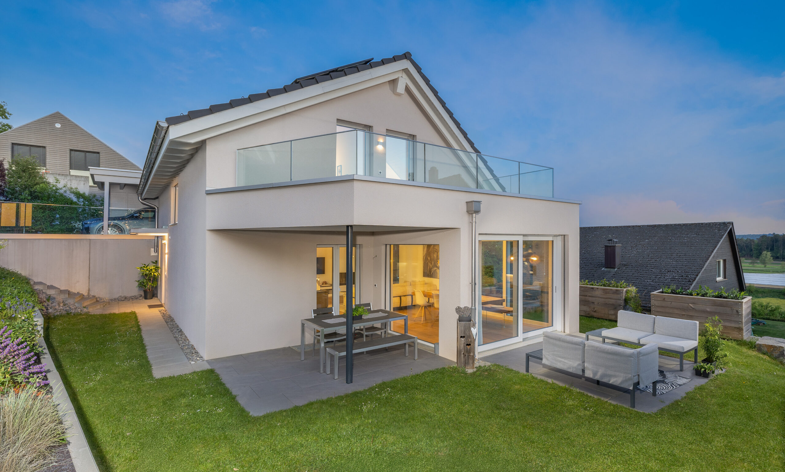 Modernes Einfamilienhaus Happy Family von Atmoshaus mit gedecktem Sitzplatz, grossem Garten und Balkon in der Abenddämmerung.